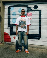 A man wearing a white graphic t-shirt with red trim, blue jeans, and orange sneakers stands in front of a white metal garage door with black graffiti.